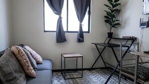 A small, compact room with a grey sofa, patterned cushions, a small table, a potted plant, and a patterned floor.