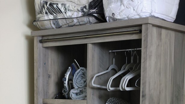 A wooden wardrobe with multiple shelves and hangers, containing folded clothes, a patterned cushion, and a rolled-up towel.