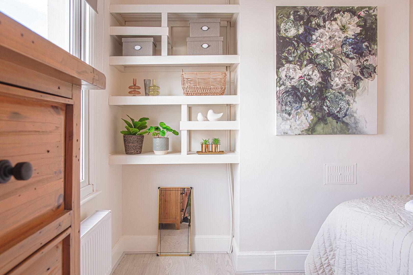 Un dormitorio con un aparador de madera, un armario con espejo, una cama blanca, un cuadro y un pequeño mueble de madera.