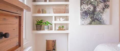 Un dormitorio con un aparador de madera, un armario con espejo, una cama blanca, un cuadro y un pequeño mueble de madera.