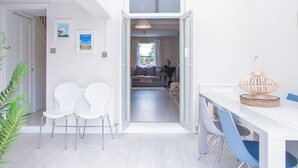 Un interno moderno con zona pranzo, sedie bianche, tavolo con decorazione intrecciata e vista su un soggiorno con divano e piante.