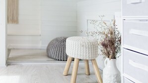 Una stanza con pavimento in legno bianco, un poggiapiedi rotondo con gambe in legno, un vaso bianco con fiori secchi e un armadietto bianco con cassetti.