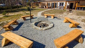 A patio area with a fire pit surrounded by wooden benches, a covered seating area, and a building with a sloped roof in the background. - Cozy apartment in Dziwnów with WiFi (Dziwnów)