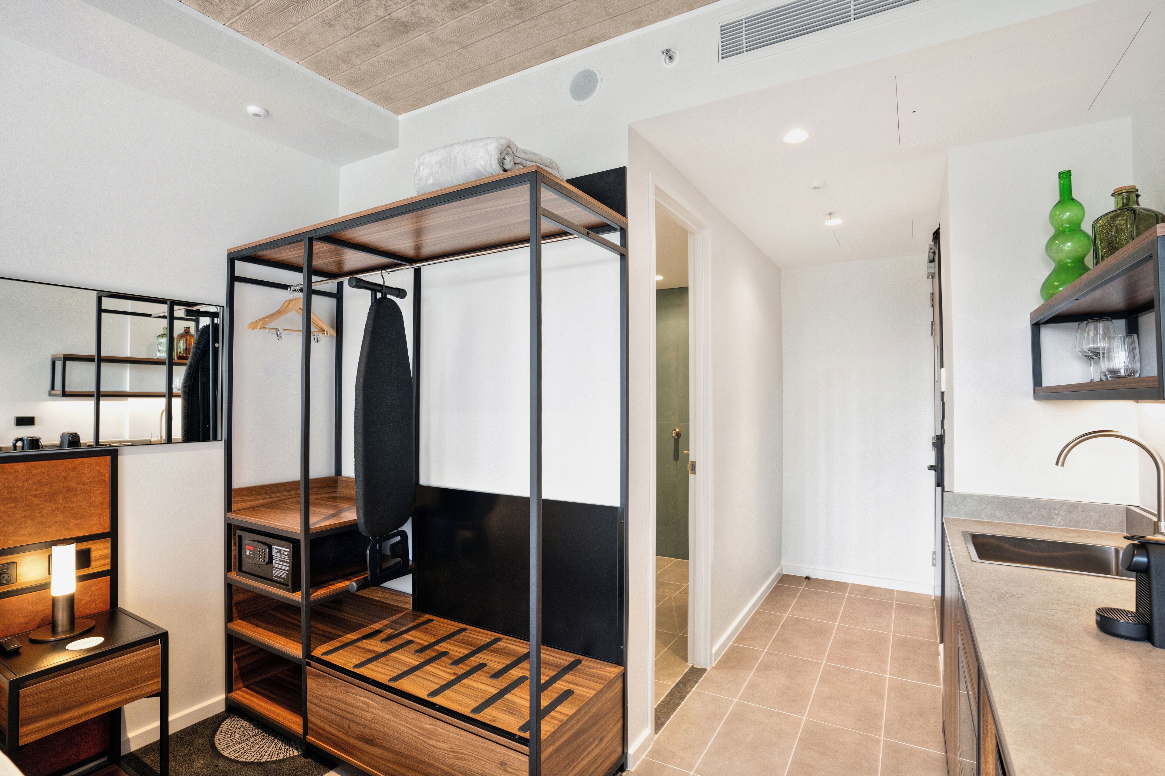 A modern hotel room with a wooden bed frame, a desk with a lamp, a bathroom with a sink, and a green bottle on the shelf.