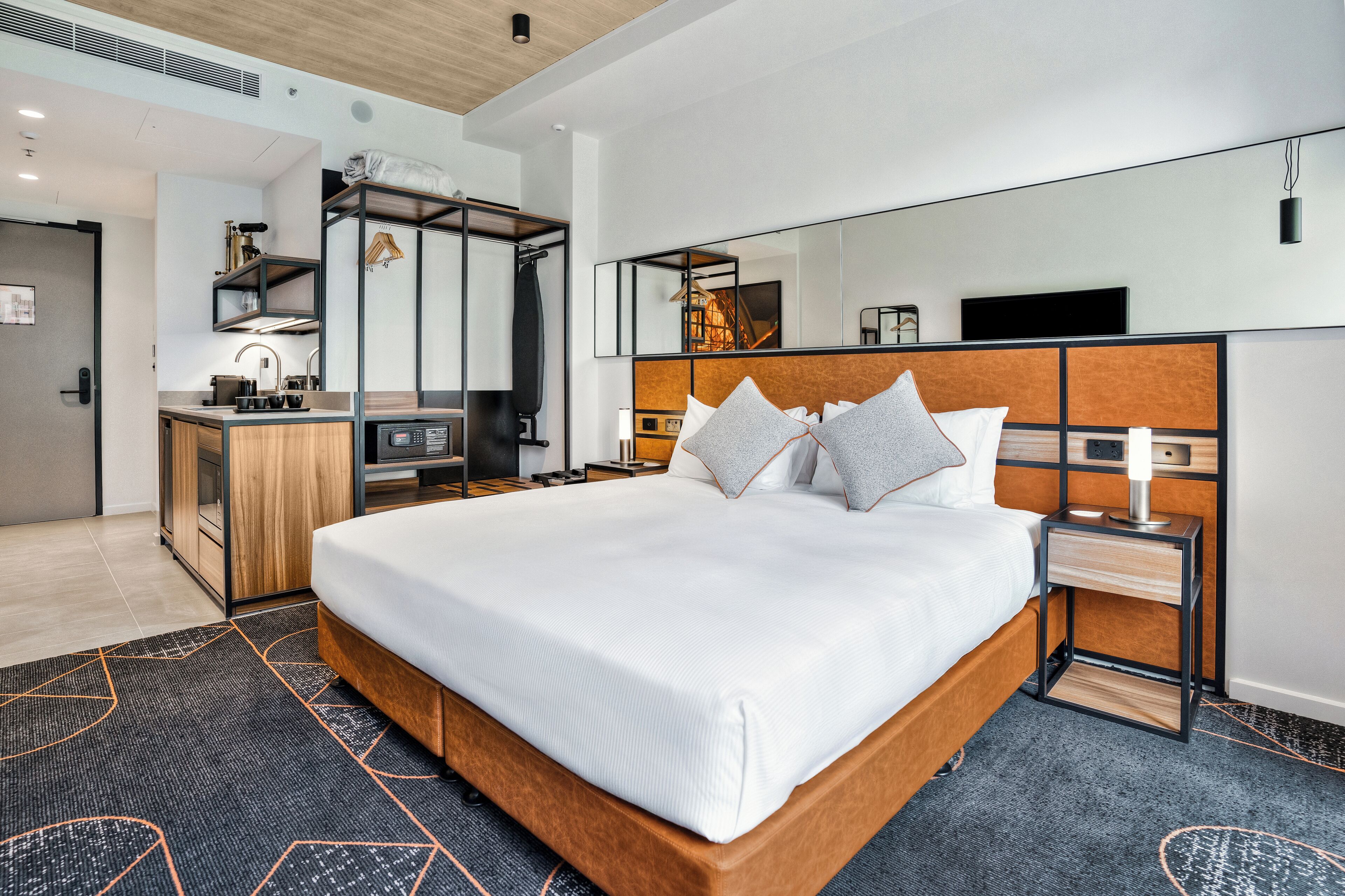 A modern hotel room with a large bed, a bedside table, a mirror, and a built-in desk with a chair.