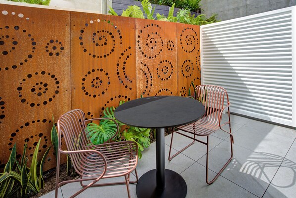 Un patio con una mesa redonda y dos sillas, un panel decorativo de metal con diseños circulares y un muro blanco con listones.