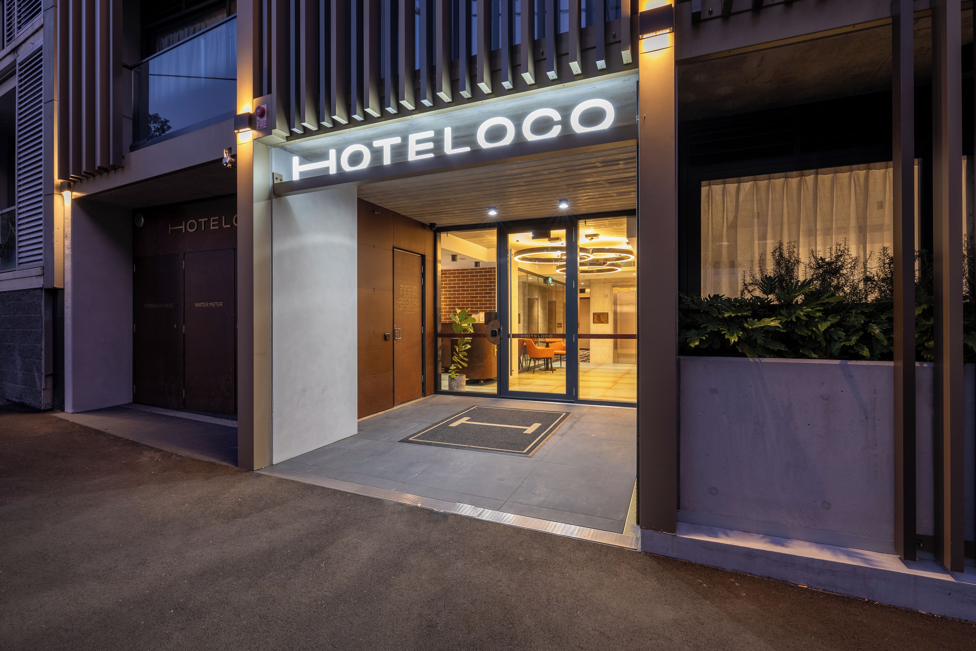 Entrada de hotel con diseño moderno, que incluye una puerta de cristal y un área de recepción visible a través del mismo.