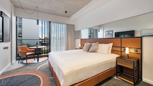 A modern hotel room with a large bed, a bedside table with a lamp, a chair, and a view of the outdoors through large windows. - Hoteloco (Alexandria)