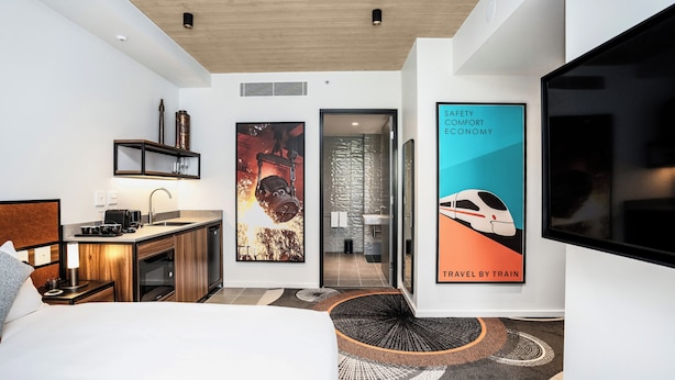 A modern hotel room with a bed, a desk, a kitchenette, and a bathroom. There are posters on the walls and a TV mounted on the wall. - Hoteloco (Alexandria)