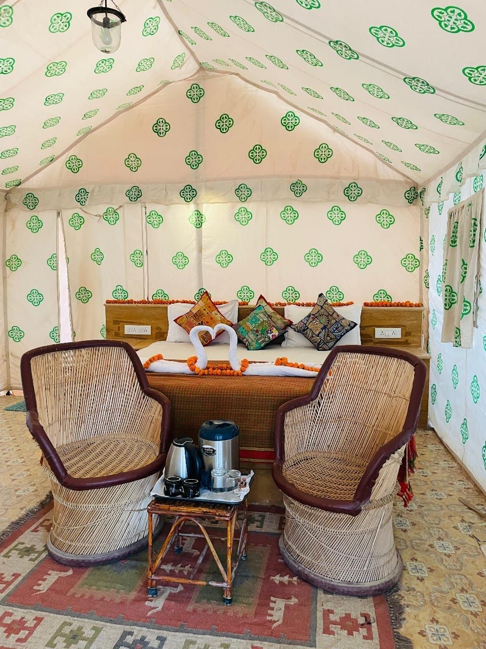 A tented room with a bed, two wicker chairs, a small table with a kettle and cups, and patterned curtains.