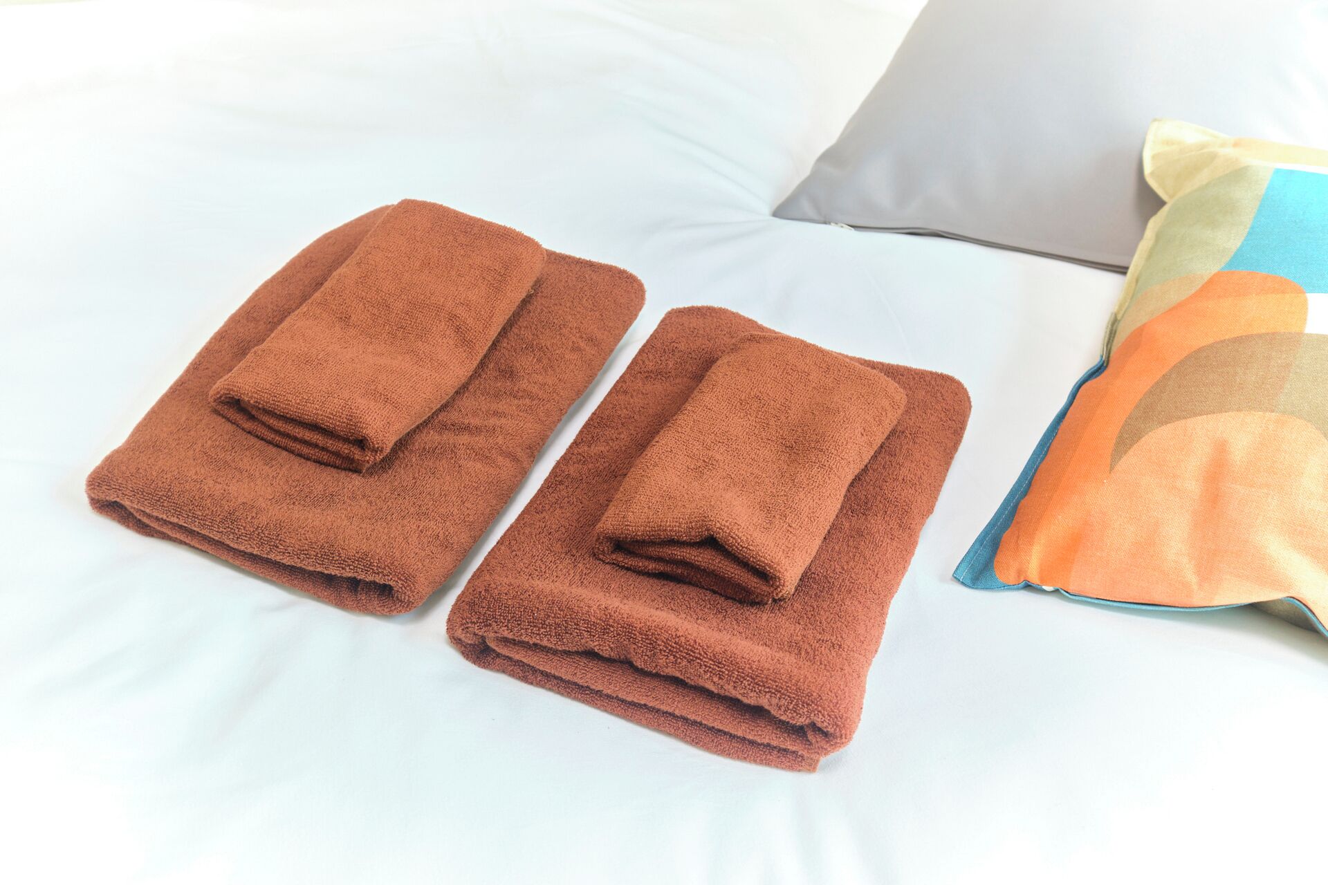Two neatly folded brown towels on a bed with white sheets and a pillow with a multicolored pattern.