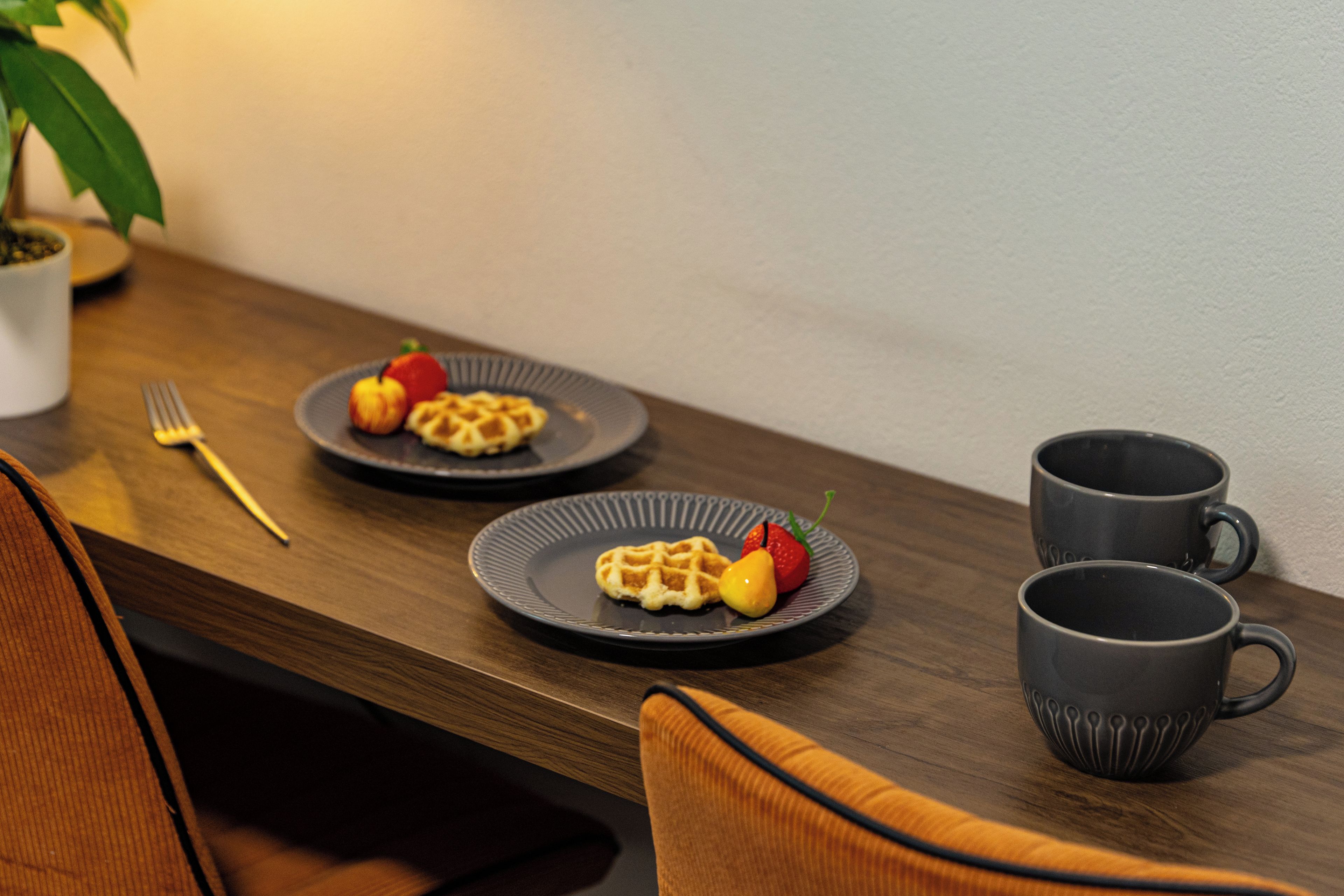 A dining table with two plates of waffles and fruit, two dark mugs, a fork, and a chair.
