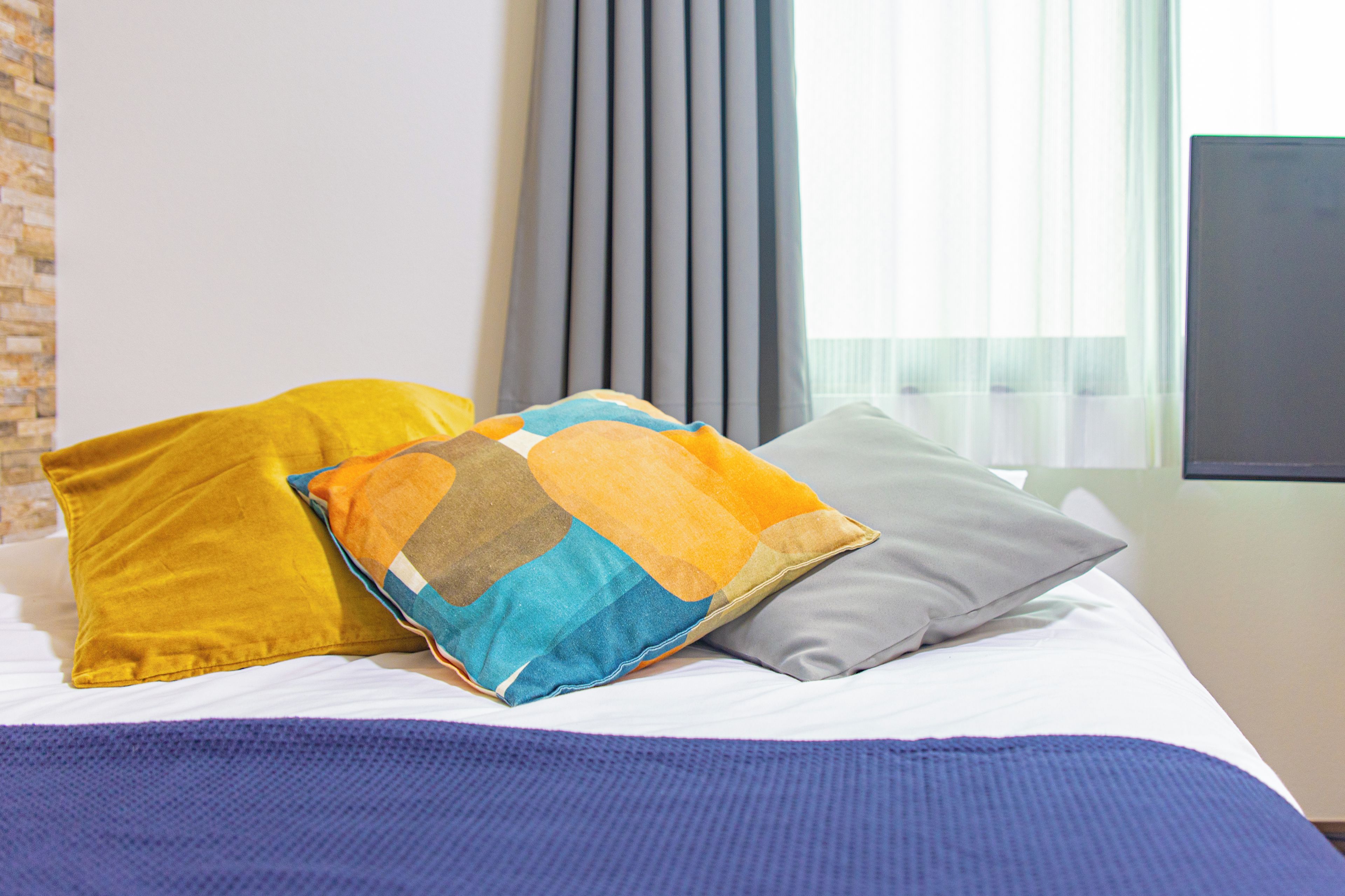 A bed with a blue blanket, two pillows in yellow and gray, and a window with blinds.