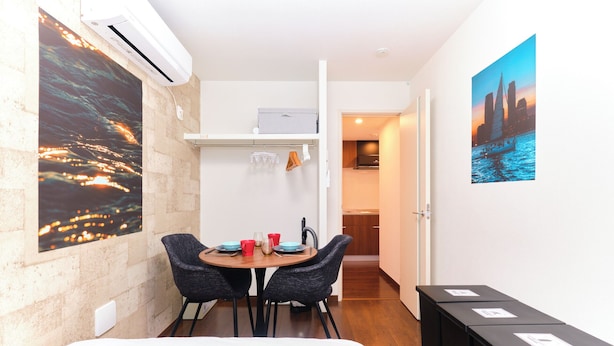 A compact hotel room with a bed, a dining table with chairs, a kitchen area, and a cityscape painting on the wall. - Le Cocon Nakarokugo by Tranova (Tokyo)
