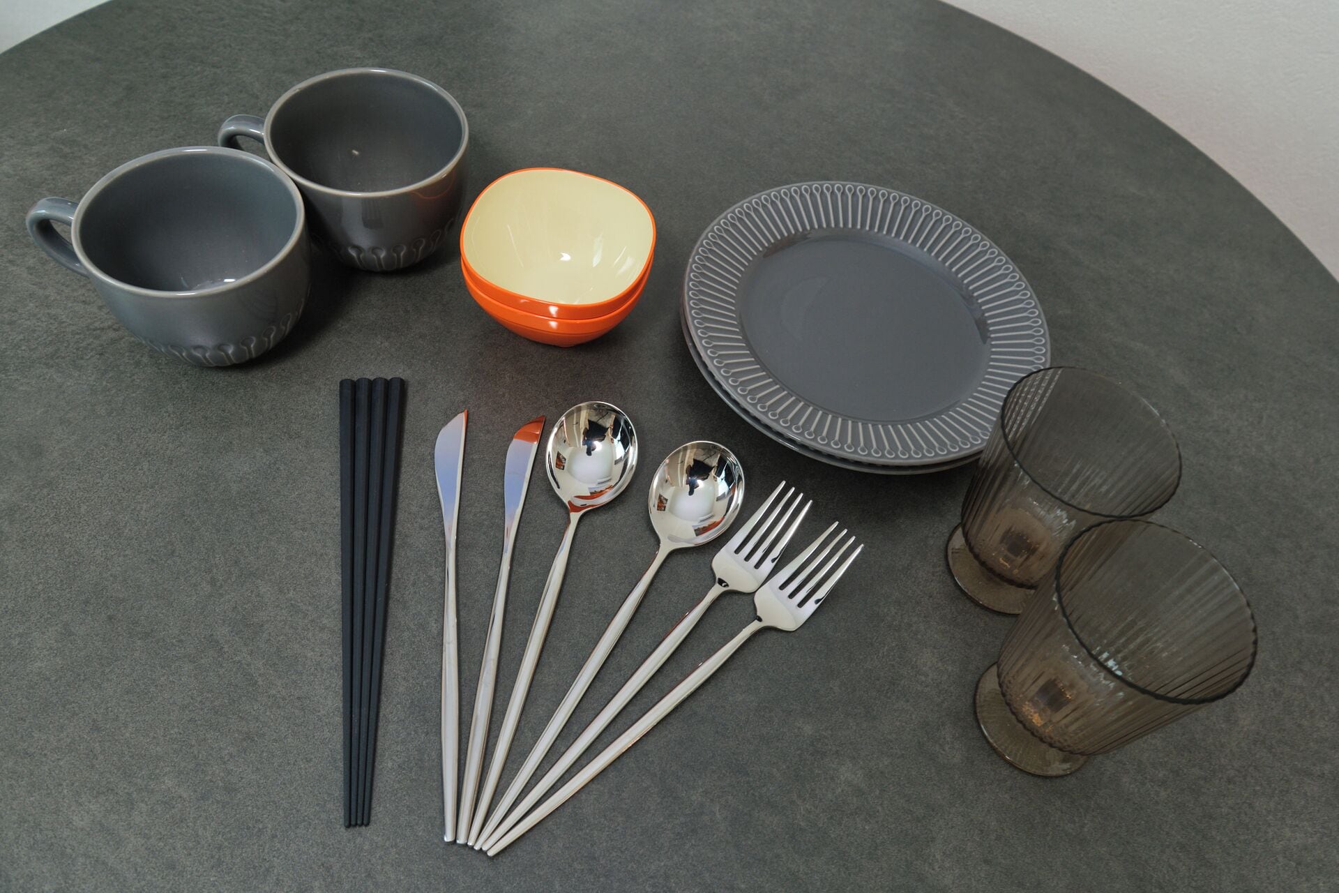 A table set with three gray cups, an orange bowl, a gray plate, four spoons, four forks, four chopsticks, and two glasses.