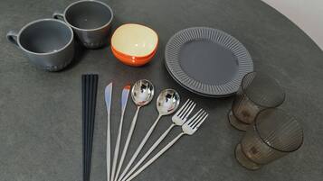 A table set with three gray cups, an orange bowl, a gray plate, four spoons, four forks, four chopsticks, and two glasses.