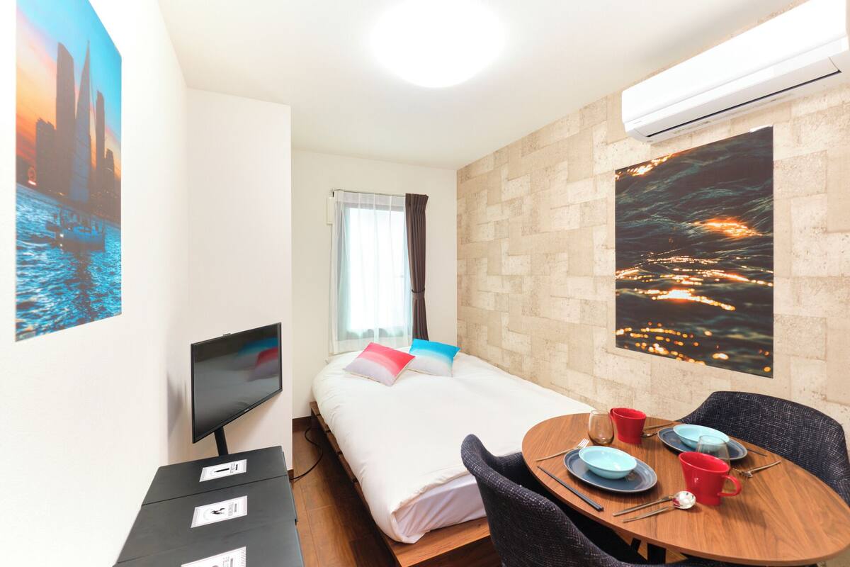 A compact hotel room with a bed, a TV, a small table with plates and cups, and a large window with a cityscape view.