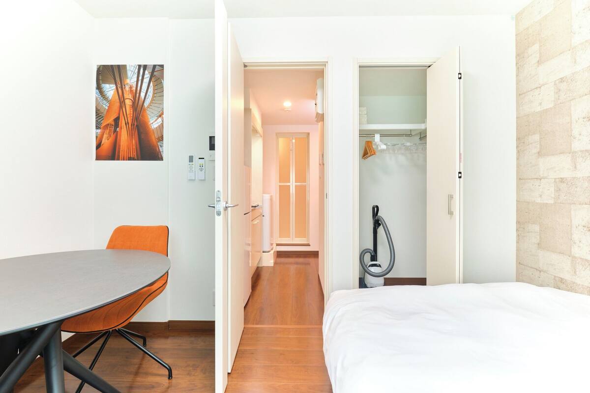 A hotel room with a bed, a desk, and a chair. There is a bathroom with a shower and a towel rack. The room has a wooden floor and a white wall.
