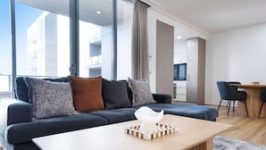A modern living room with a dark sofa, a wooden coffee table, and a view of a balcony and building through large windows.