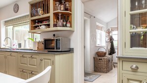 A kitchen with a microwave, shelves with bottles, a clock, and a dining area with a chair. - Beautiful 3BD Fam Retreat in Oxfordshire (Chipping Norton)