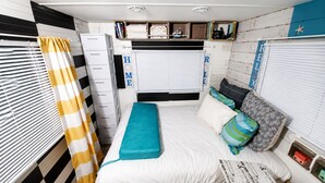 A compact bedroom with a bed, storage drawers, a window with blinds, and a small shelf with personal items. - VINTAGE RV & SPA Experience for 3 (Dover)