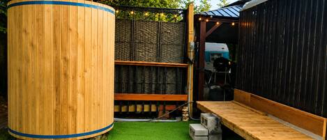 A wooden outdoor seating area with a green artificial turf, a large cylindrical object, and a black structure with a sloped roof.
