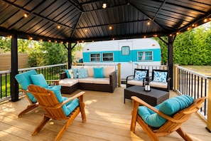 En overdækket terrasse med træmøbler og en blå campingvogn i baggrunden.