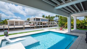 A swimming pool with a slide, a covered patio area, and a view of a waterfront property with a house and palm trees. - Big Pine Paradise: Beautiful Updated 4BR Canal Front Home, Pool, Dockage, Easy Gulf/Atlantic Access (Big Pine Key)