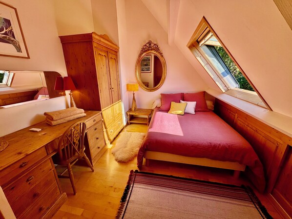 A bedroom with a wooden desk, a bed with red bedding, a mirror, and a window with a view of greenery. - Nid Douillet Les Mouettes, vue sur mer (Fouesnant)
