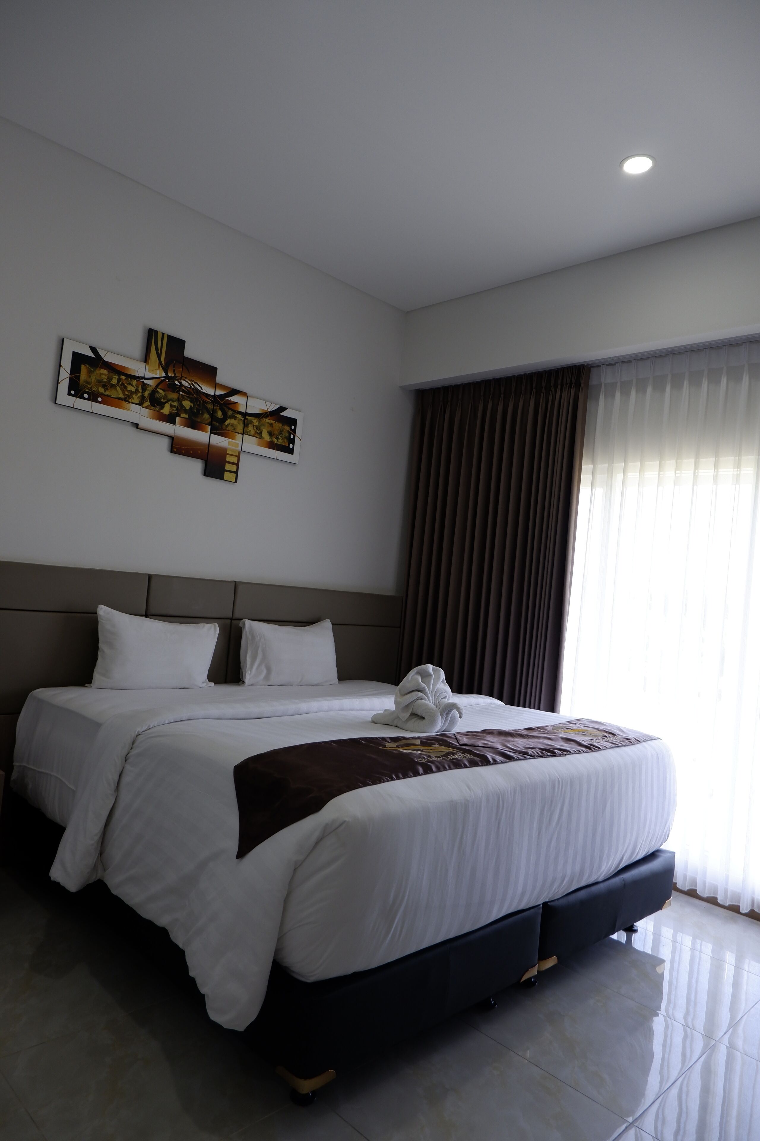 A hotel room with a large bed, a headboard, a wall-mounted artwork, and a window with curtains.
