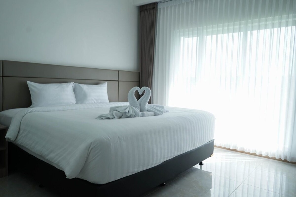 A modern hotel room with a large bed, a headboard, and sheer curtains.