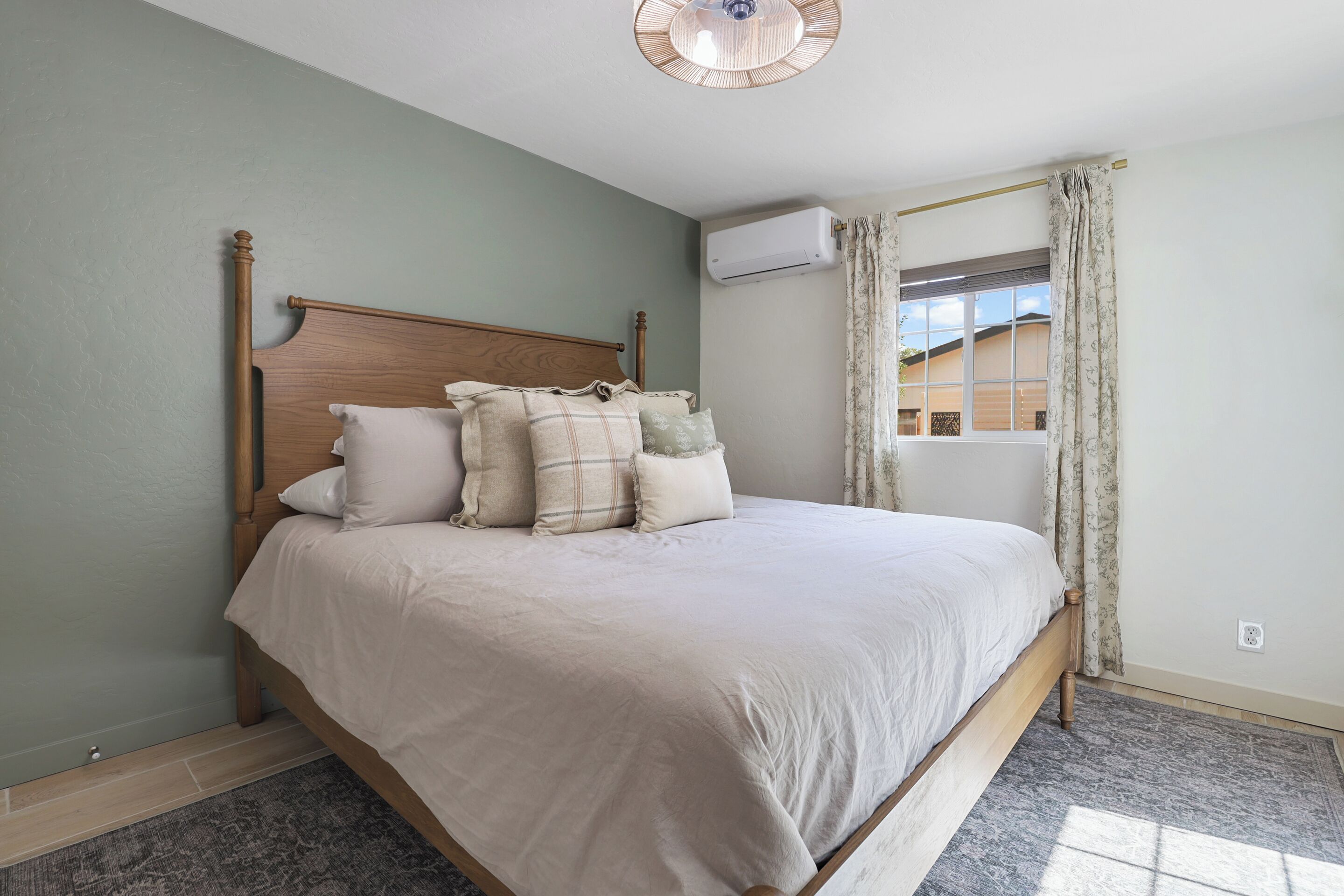 A neatly made bed with a wooden headboard, a window with curtains, and an air conditioning unit on the wall.