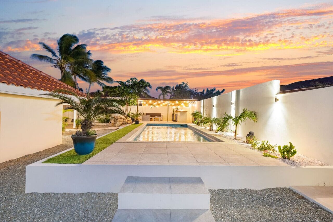 A modern outdoor pool area with a clear view of the sky, surrounded by palm trees and a well-maintained lawn.