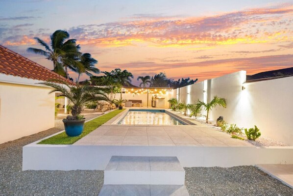 A modern outdoor pool area with a clear view of the sky, surrounded by palm trees and a well-maintained lawn.