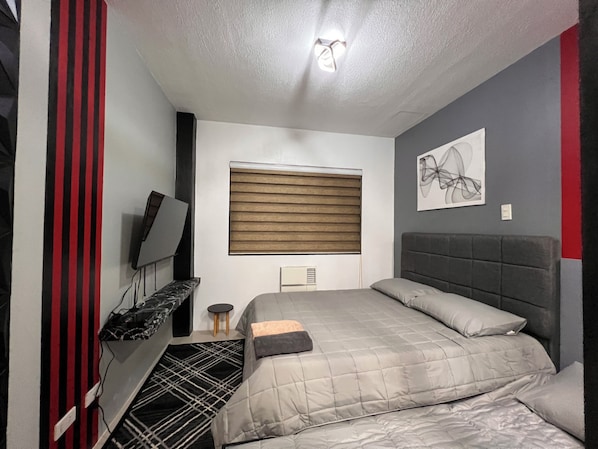 A modern bedroom with a bed, a TV, a small stool, and a window with blinds. - Hubomoto People's Suite (Iloilo)
