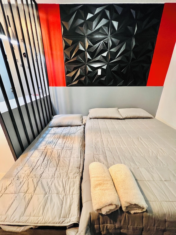 A double bed with a grey quilt, two pillows, and two rolled towels. The headboard has a geometric pattern, and there's a red curtain on the left.