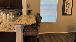 A kitchen with dark wooden cabinets, a granite countertop, a wicker chair, a window with blinds, and a framed artwork on the wall. - Modern Cozy Retreat in Katy, TX (Katy)