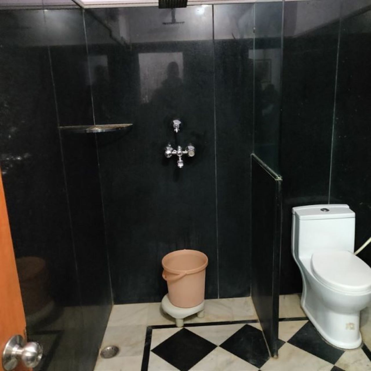 Ein Badezimmer mit schwarzem Fliesenbelag, einer weißen Toilette, einem pinkfarbenen Mülleimer und einer Duschkabine aus Glas.
