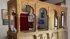A carved wooden structure with intricate designs, including figures and columns, in a room with a ceiling fan and framed pictures on the walls. - Hotel Bharat Villas Jaisalmer (Jaisalmer)