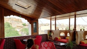 A covered outdoor seating area with red chairs and a table, overlooking a cityscape. - Hotel Bharat Villas Jaisalmer (Jaisalmer)