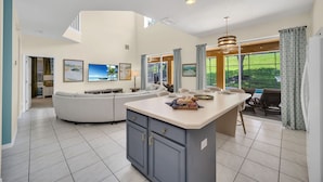 A spacious living area with a curved sofa, a kitchen island, and a dining area with a view of the outdoors. - Spacious Villa: Pool, Game Room & Themed Rooms (Kissimmee)