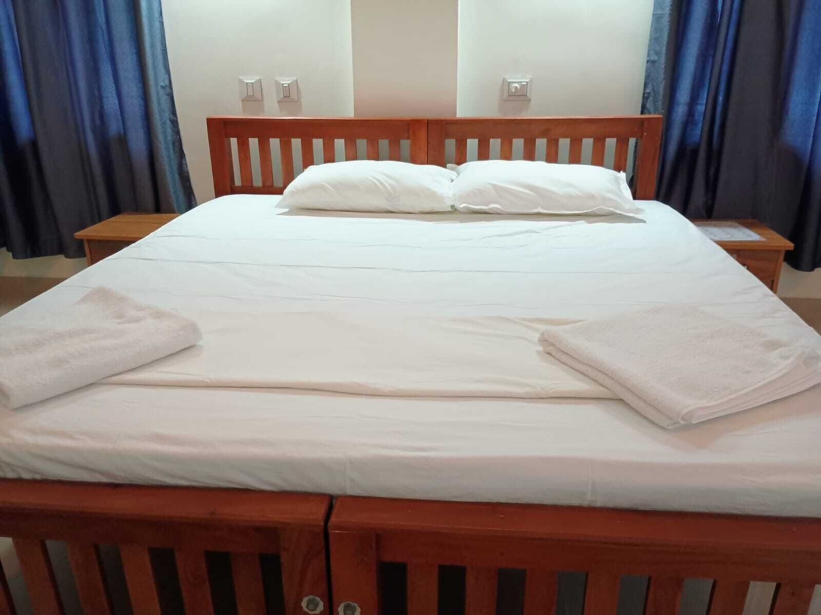 Uma cama bem arrumada com lençóis e almofadas brancos, uma cabeceira de madeira e duas mesas de cabeceira com abajures.