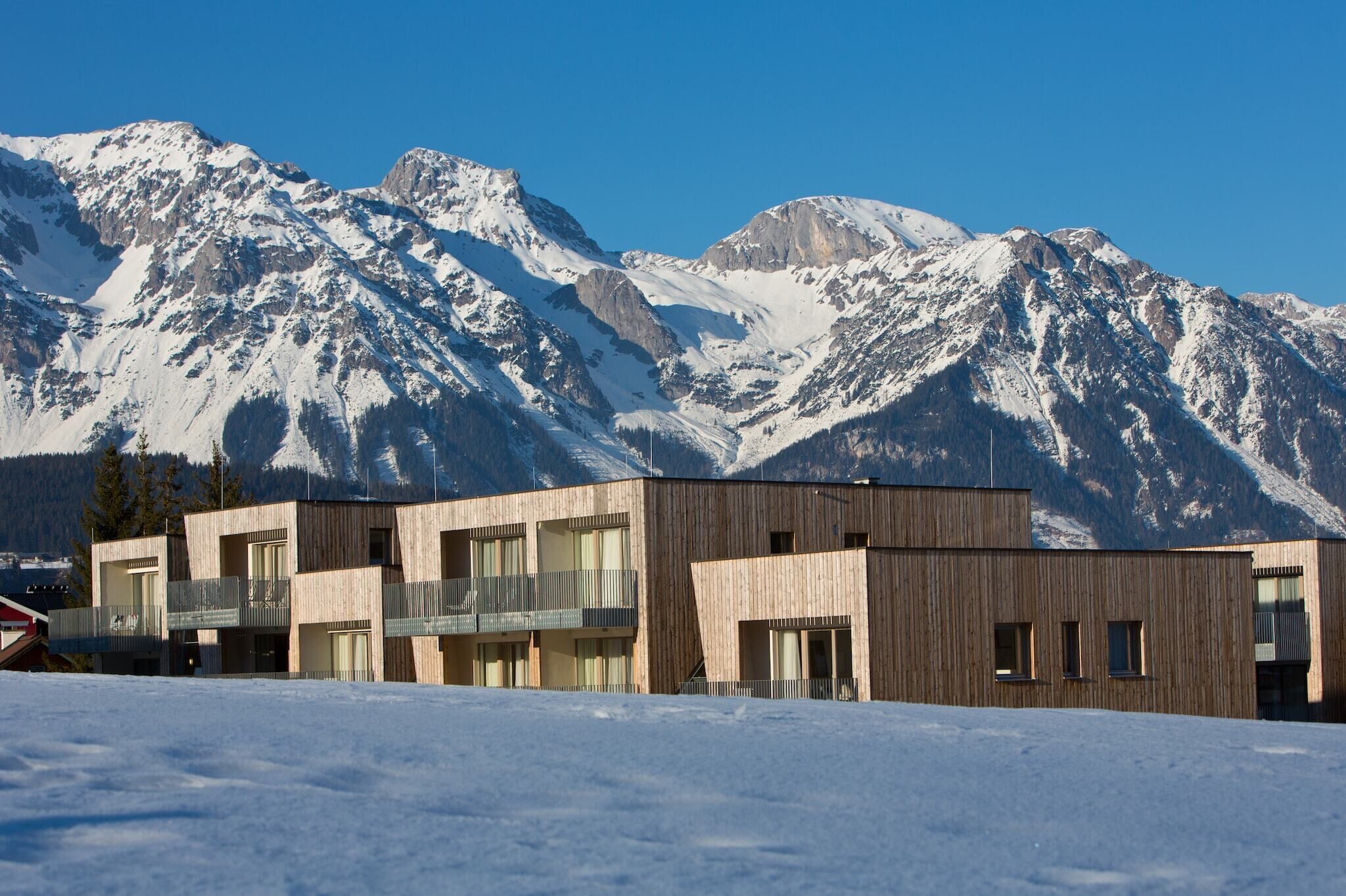 Un bâtiment moderne à la façade en bois, doté de nombreux balcons et de grandes fenêtres, qui se détache sur un paysage de montagnes enneigées.