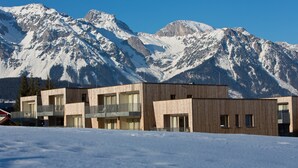 A modern building with a wooden facade, multiple balconies, and large windows, set against a backdrop of snow-covered mountains. - Lively apartment near the ski slope (Schladming - Rohrmoos)