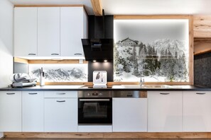 Une cuisine moderne avec des armoires blanches, un four encastré, une hotte noire et une grande fenêtre donnant sur un paysage de montagne enneigé.