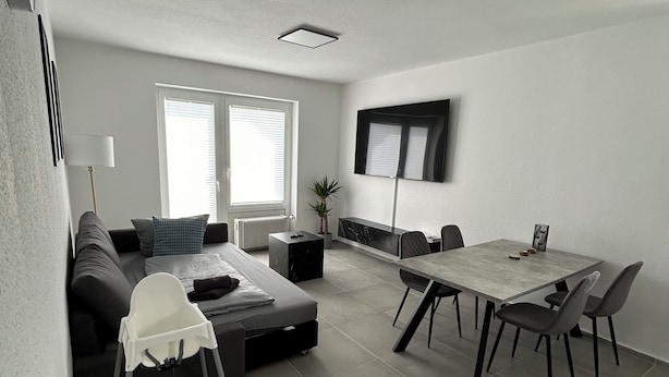 A modern living room with a sofa, a dining table with chairs, a TV mounted on the wall, and a window with blinds. - Vacation apartment Menges (Medebach)