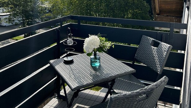 A balcony with wicker furniture, a table with a vase of flowers, and a view of trees and buildings. - Ferienwohnung Juli - neu Renovierte, Moderne Ferienwohnung (Medebach)