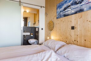 Une chambre avec deux lits, un mur en bois, un tableau, et une salle de bain visible à travers une porte ouverte.