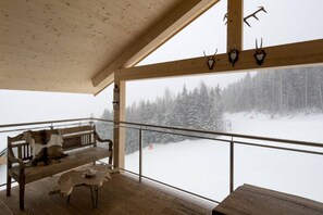 Een houten terras met een bank en tafel, met uitzicht op een besneeuwd landschap met bomen.