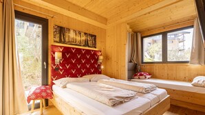 Une chambre dans une cabane en bois, avec un grand lit, un banc muni d’un coussin rouge, un tableau accroché au mur et une fenêtre avec des rideaux.